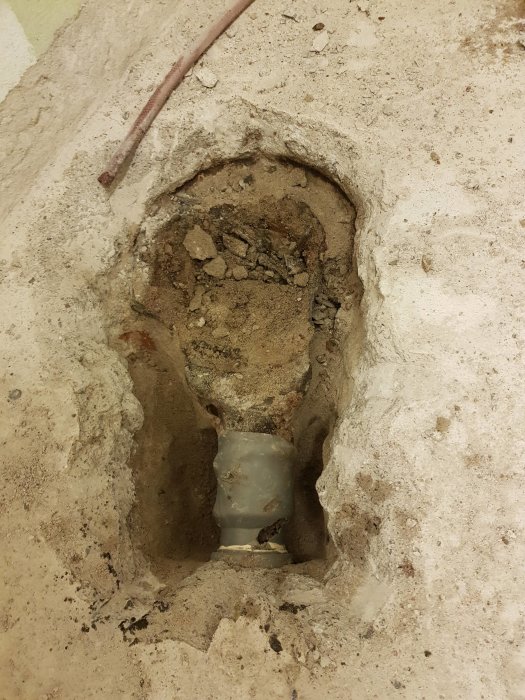 Ett plaströr upptäckt i ett utgrävt hål i ett betonggolv, med synliga omgivande stenar och grus.