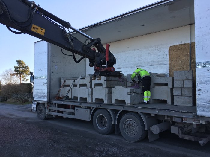 Lastbil med kran lossar Vertiblock-stödmurar på byggarbetsplats, med arbetare som säkerställer lasten.