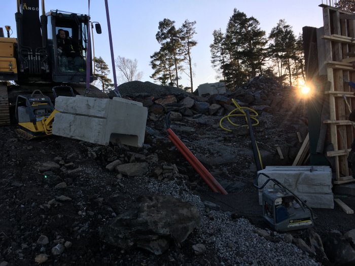 Byggarbetsplats med Vertiblock, maskiner och byggmaterial, och en grävmaskinförare i förgrunden.