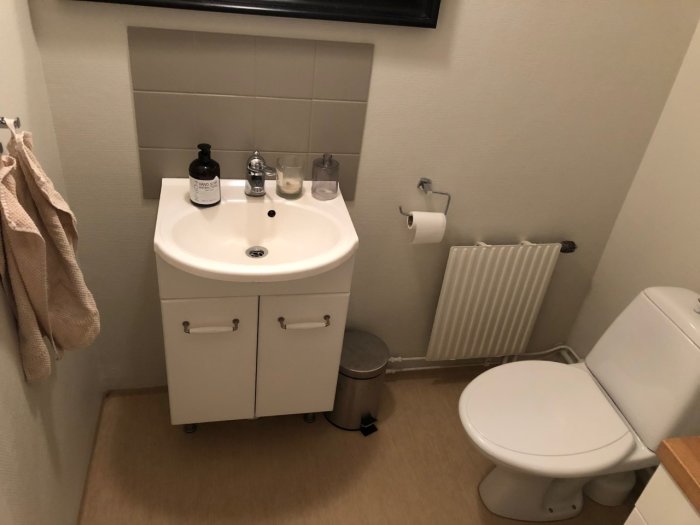 Ett litet badrum med vit tvättställsskåp, toalett, radiator, kakelväggdelar, och vävklädda väggar redo för renovering.