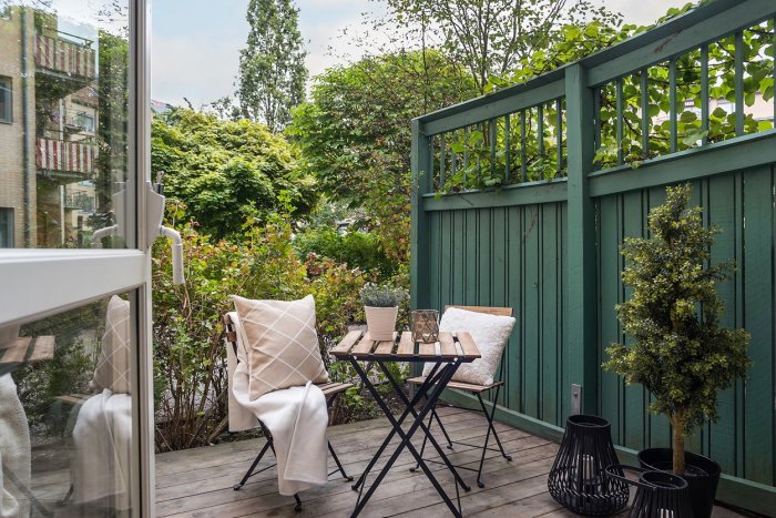 Balkong med trästaket, gröna buskar, ett litet bord med två stolar och dekorativa föremål, under sommartid.