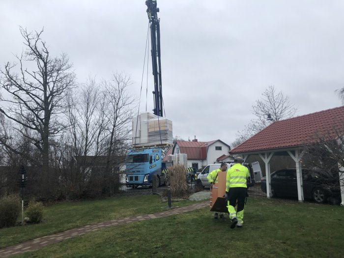 Kranbil levererar byggmaterial medan två arbetare bär en kartong på en bostadstomt.