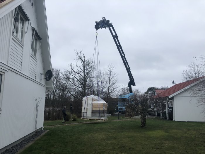 Kran lyfter stor leverans nära vit huskropp, några personer och en lastbil i bakgrunden.