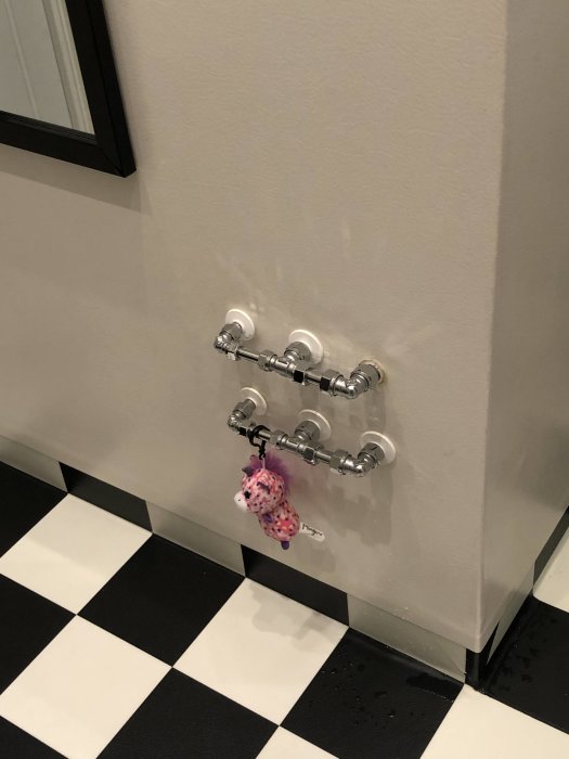 Väggmonterad kroklist med fyra krokar och en hängande rosa leksak i ett badrum med schackrutigt golv.