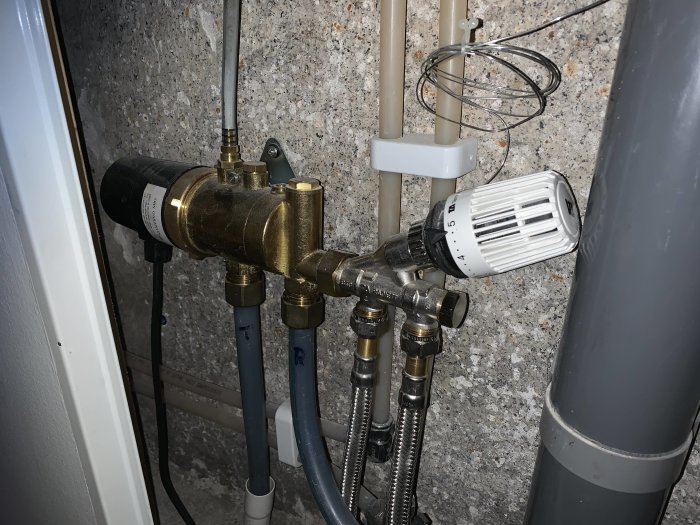 Vattenburet golvvärmesystem med termostat, kopplingar och en okänd cylinderformad enhet i en källare.