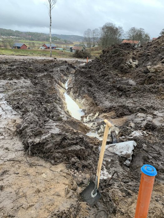 Grävd ränna fylld med vatten för makadam och återfyllning, spade i förgrunden, byggprojekt på landsbygd.