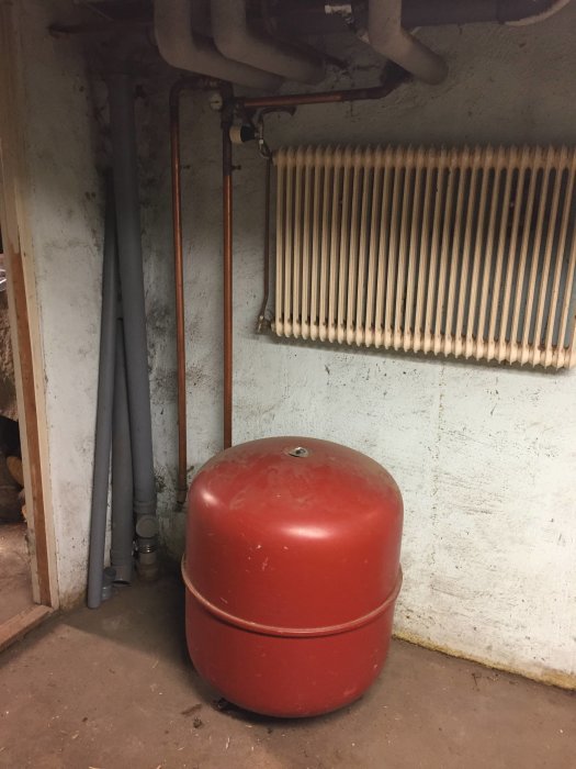 Expansionskärl i rött placerat i ett pannrum, intill ett värmeelement och rörledningar.