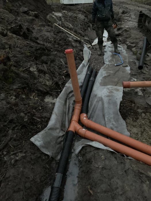 En person leder rörarbete för dränering under en gruslagd väg med olika dräneringsslingor synliga vid skymningen.