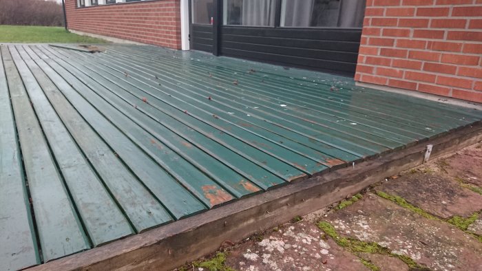 Gamla och slitna träplankor på ett trädäck med synliga tegelstenar och gräsmatta i bakgrunden.