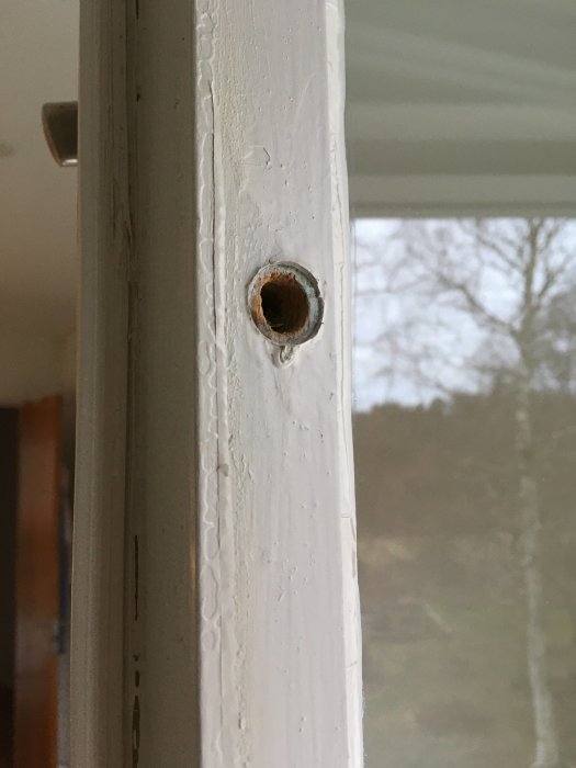 Närbild på ett fönsters skruvhål med en lossnad hullingshylsa, vitmålad ram mot suddig bakgrund.