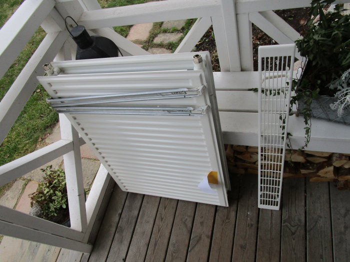 Gulvit sönderfrusen panelradiator av okänt märke och ett toppgaller på träaltan.