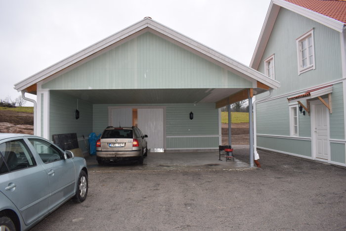 Carport med bil och hus i ljusgrön färg med vita knutar och röd takpannor, med byggmaterial vid sidan.