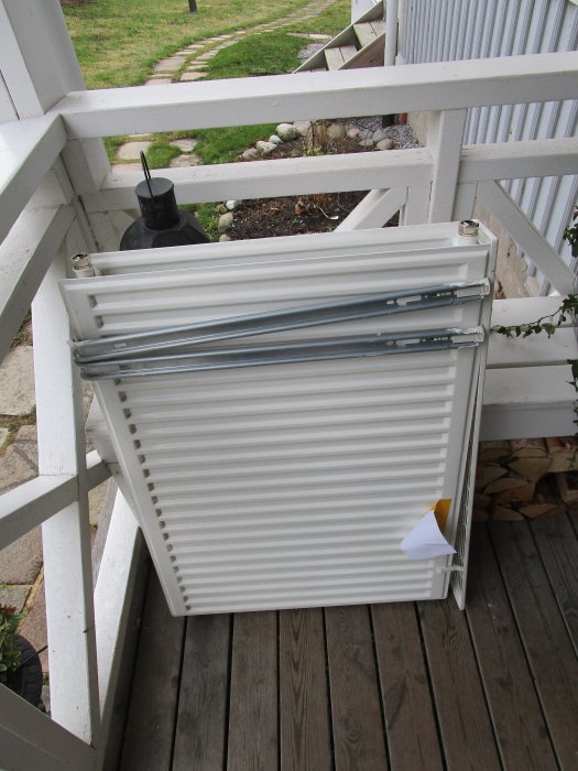 Vit panelradiator står på träaltan med trädgårdsutsikt, efterfrågas för identifiering.