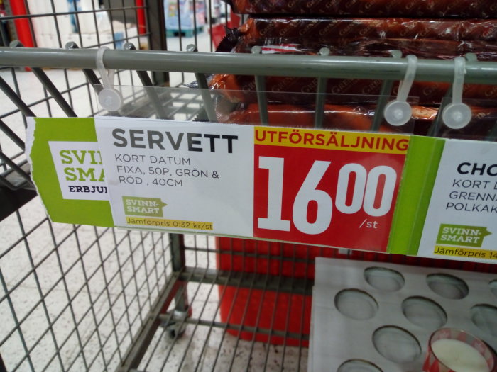 Rea-skylt där det står "SERVETT KORT DATUM FIXA, 50p, GRÖN & RÖD, 40CM" med pris "16:00" i en shoppingkorg.