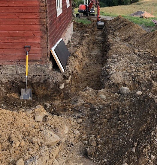 Grävning för dagvattenledning nära husgrund med rödfärgat trähus, spade och grävmaskin i bakgrunden.