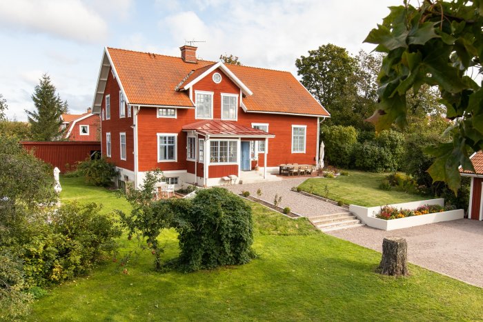 Rödmålat traditionellt hus med två våningar, vita fönster, grönt gräs och anlagd trädgård.