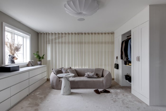 Elegant sovrum med beige soffa, inbyggda garderober, vit fåtölj och designertaklampa.