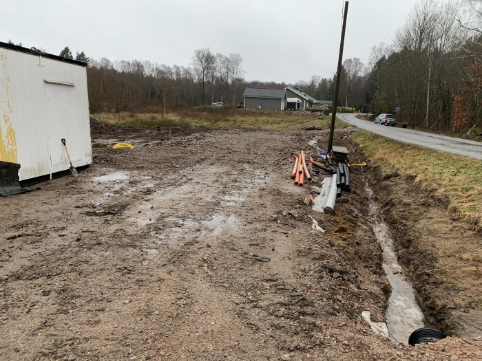 Pågående dräneringsarbete med utlagt byggmaterial längs en lerig byggplats bredvid en väg.