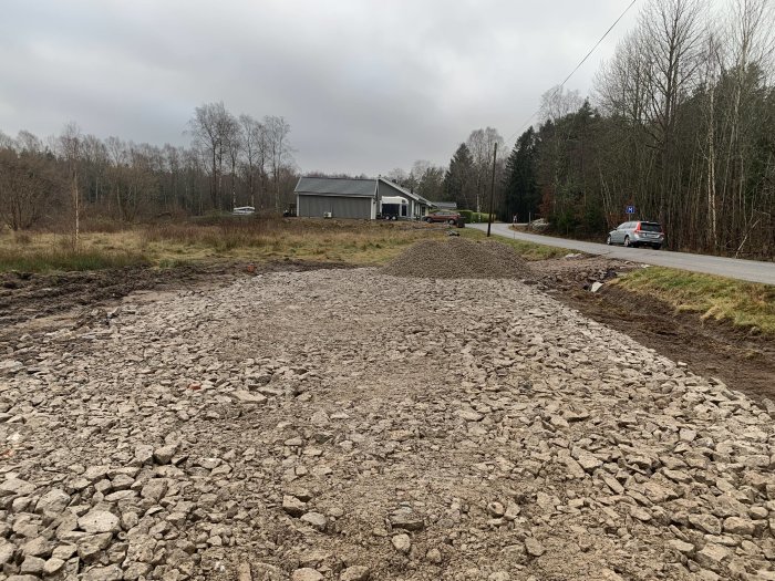 Nyplanerad yta med lager av grova sten 0-150 för dränering vid infart med skog i bakgrunden.