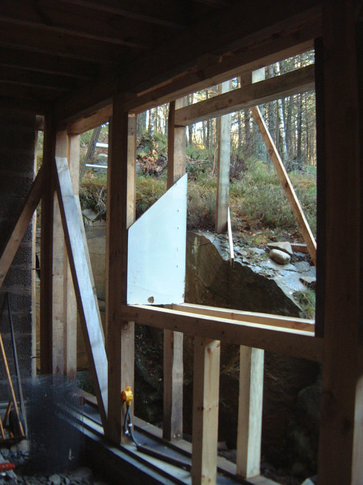 Inre stommen i ett pågående byggprojekt med dubbla träreglar för isolering, utsikt till skog och gård.