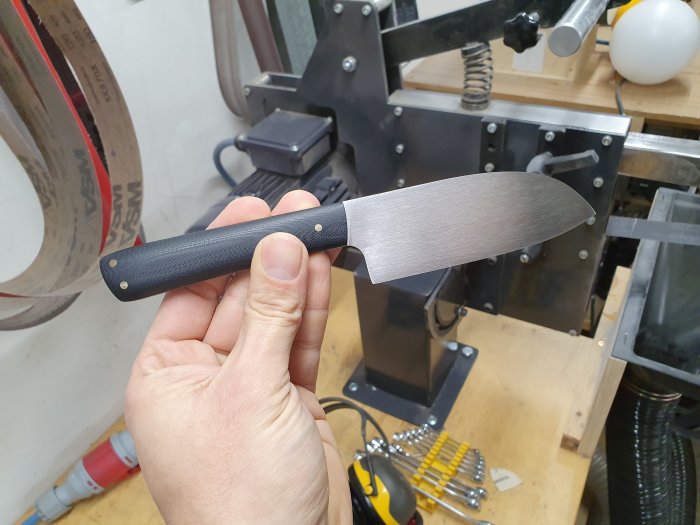 Hemgjord grönsakskniv som liknar en santoku, med RWL-34-blad och G10-skaft, på en verkstadsbänk.