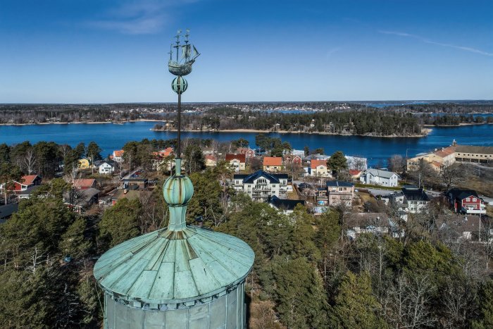 Utsikt över Vaxholm med hus, träd och vatten från ett torns spira mot en klarblå himmel.