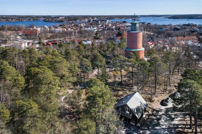 Flygbild över Vaxholm med gammalt torn och hus omgivna av skog och hav i bakgrunden.