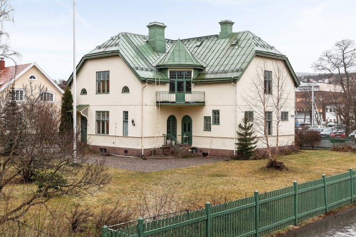 Välbevarad äldre villa med grönt tak och balkong, omgiven av trädgård och staket.