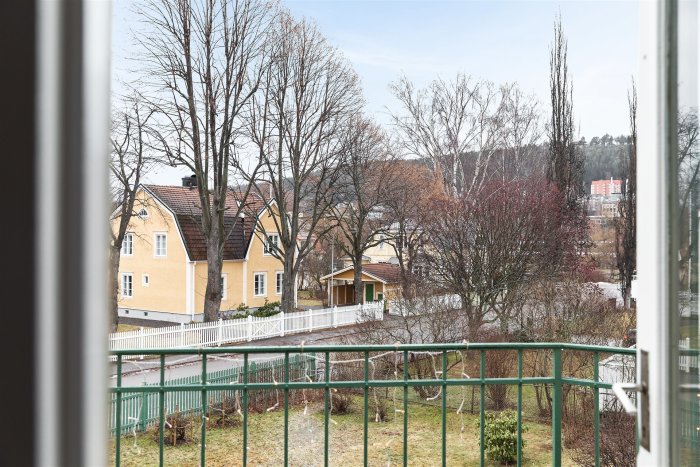 Vy från fönster av bostadsområde med gula hus, träd och ett grönt staket.