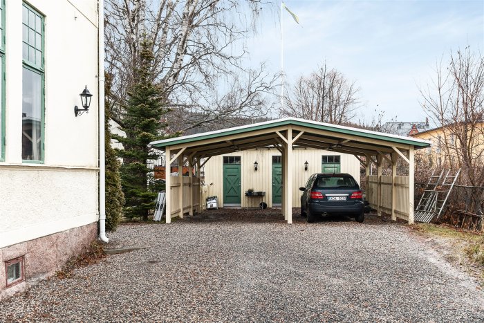 Bil parkerad under ett trädäckande carport intill ett traditionellt hus.