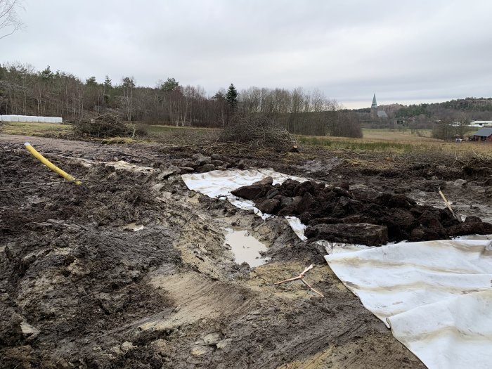 En pågående vägarbetsplats med lerig mark, ett gult 110-elrör och vita geotextiler, med natur och en kyrktorn i bakgrunden.