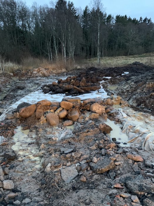 Grävningsarbete för vägbygge med synliga stenar, jord och ispölar i skymningsljus.