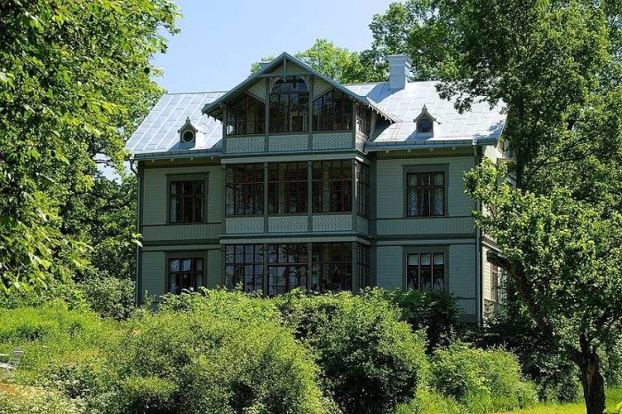 Traditionellt grönmålat trähus med veranda och skiffer tak omgivet av grönska under en klar dag.