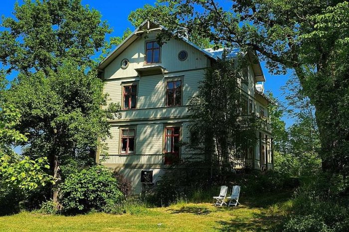 Trehus med flera våningar, målat i pudrigt blått bland grönskande träd och buskar, med två vita trädgårdsstolar framför.