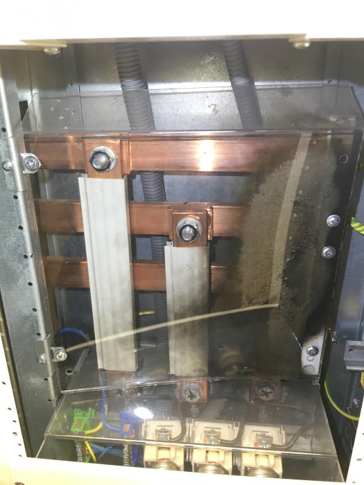 Elektriskt skåp med kopparskenor och genomskinligt beröringsskydd med sotspår och felmonterade skruvar.