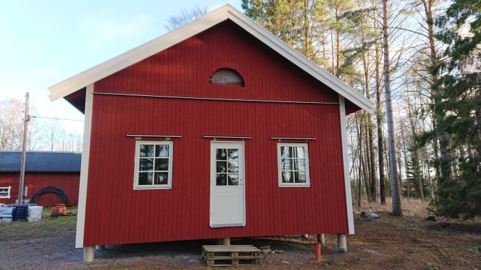 Nybyggt rött hus med nysatta vita fönsterfoder och dörr, huset omgivet av skog.