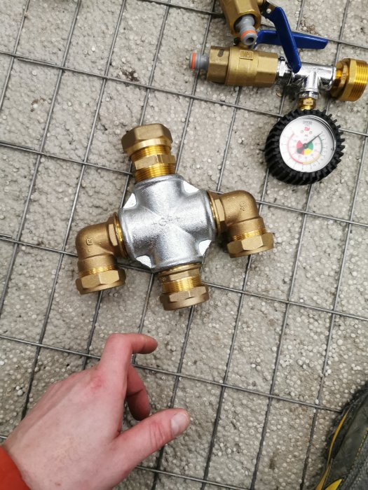 Hand vid en kraftig metallisk korsformad ventil med anslutningar, placerad på ett kaklat golv.