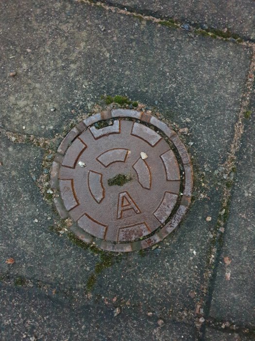 Rostig brunnslock av metall med bokstaven A i mitten, inbäddat i ett stenbelagt golv med mossa.