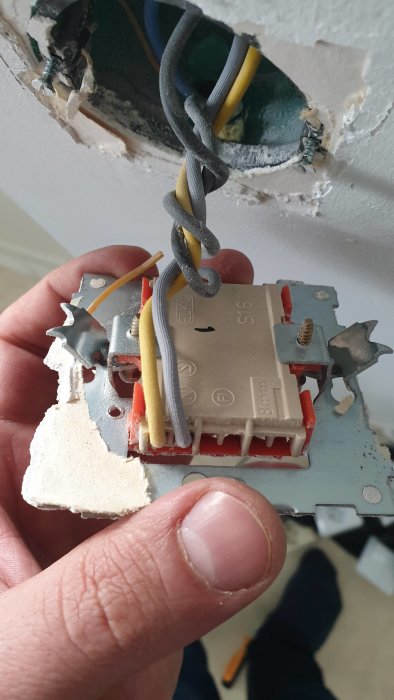 Hand som håller en delvis installerad eluttag mot en vägg med exponerade elledningar och en skadad gipsvägg.