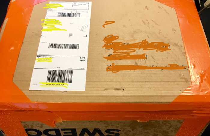 Ett slitet och skadat paket med övertäckt fraktetikett och orange packtejp, tecken på bristande omsorg vid förpackning.