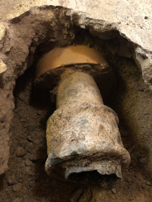 Gammal röranslutning i betong med en bit av rostigt gjutjärnsrör som sticker ut, sedd i en grävd öppning.