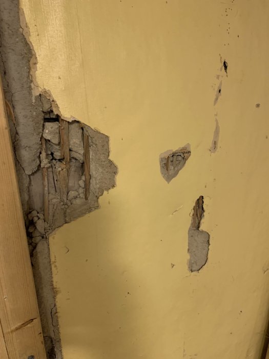 Skadad gul vägg med lagningar och puts som fallit bort, exponerar underliggande vass och ståltråd.