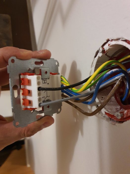 En hand håller en Vadsbo UDN100 universal-pushdimmer framför en öppen väggdosa med synliga elkablar.