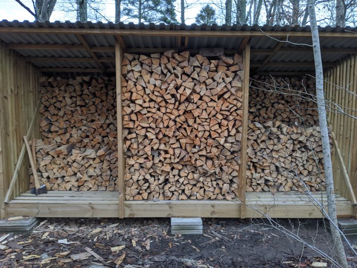 Vedträn staplade i vedbod med yxa framför, rester av klyvning synliga på marken.