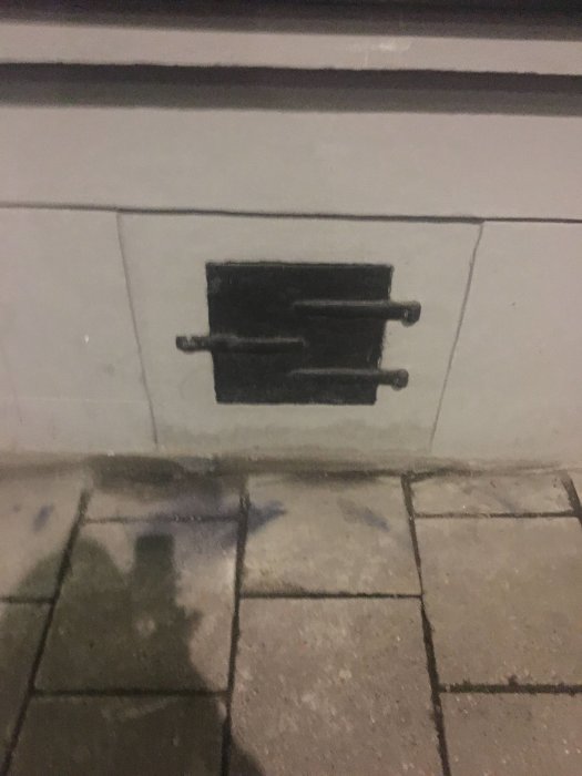 Ett litet svart källarfönster med galler stängt, inbyggt i en vitputsad vägg över stenplattor, nattetid.