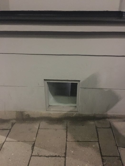 En källarfönsterlucka på ett gathus med vit fasad och trottoar i förgrunden.