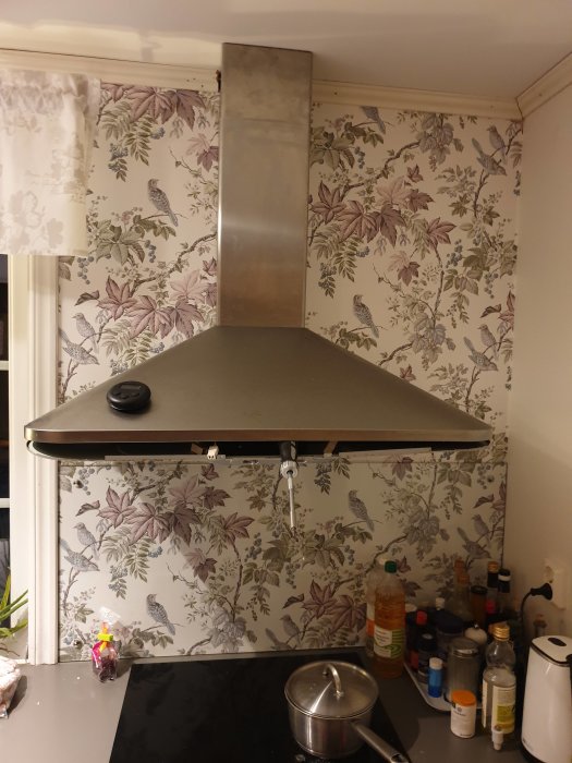 Kök med äldre köksfläkt från IKEA, utan fungerande belysning, monterad över spis och blommig tapet i bakgrunden.