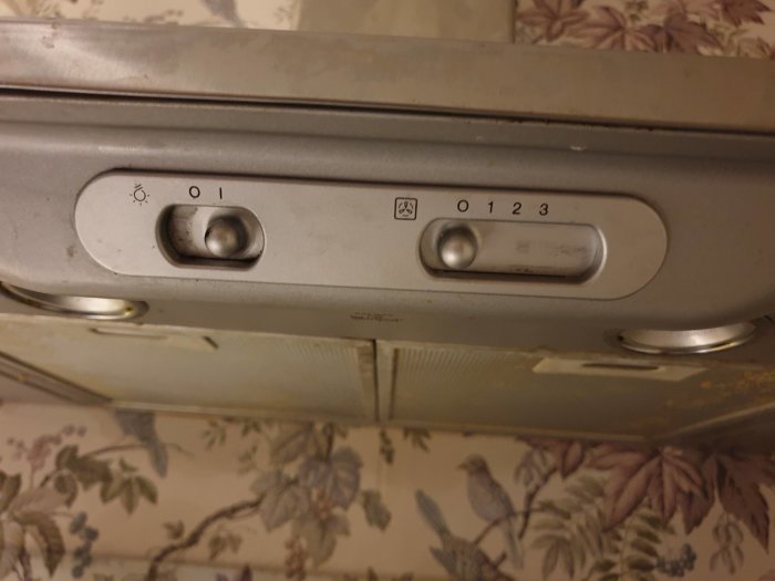 Gamla fläktreglage med vred för strömbrytare och fläkthastigheter mot blommigt tapetbakgrund.