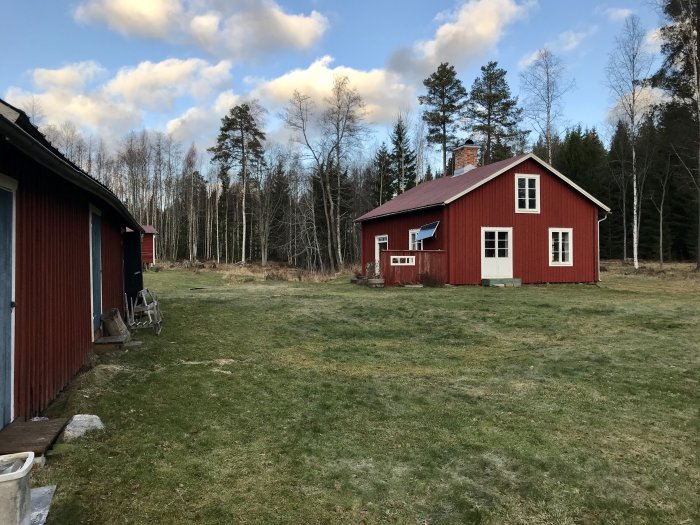 Rödmålat gammalt torp med vita knutar i lantlig miljö, bredvid skogskant.