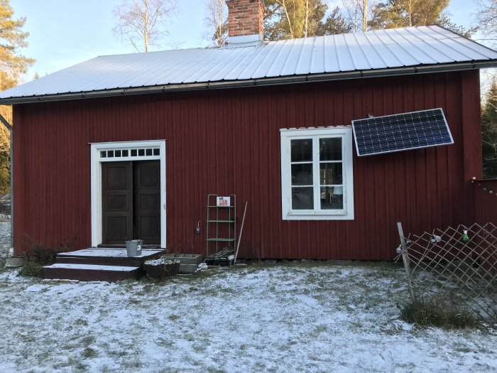 Ett rött torp från 1800-talet med vita knutar, en solpanel och snö på marken under en solig dag.